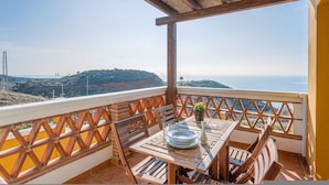 A balcony with a wooden table and chairs, a potted plant, and a view of the sea and hills. - Villa Margarita Villa Margarita Solset (Velez-Malaga)