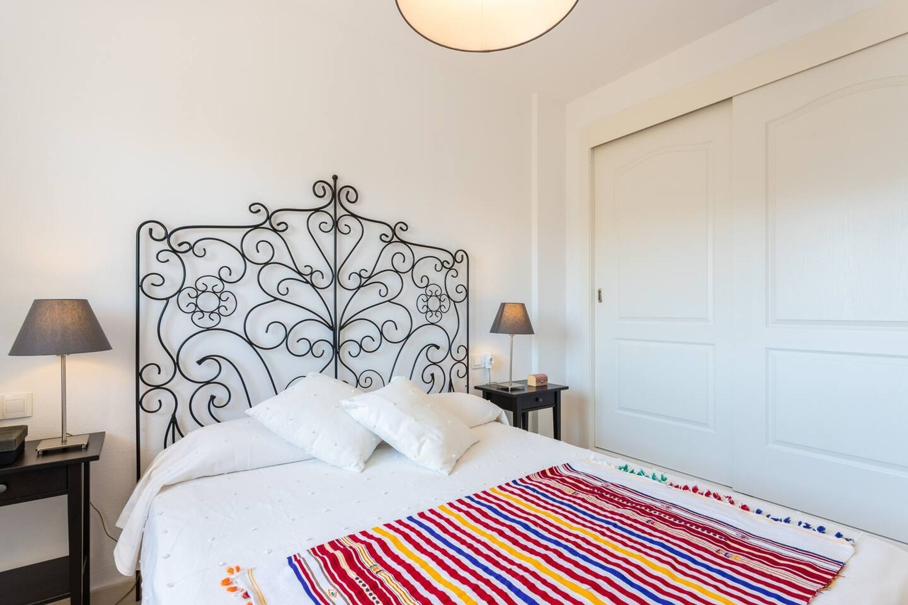 Een net opgemaakt bed met een witte dekbedovertrek en een kleurrijke gestreepte deken, een zwart metalen hoofdbord, twee nachtkastjes met lampen, en een witte deur op de achtergrond.