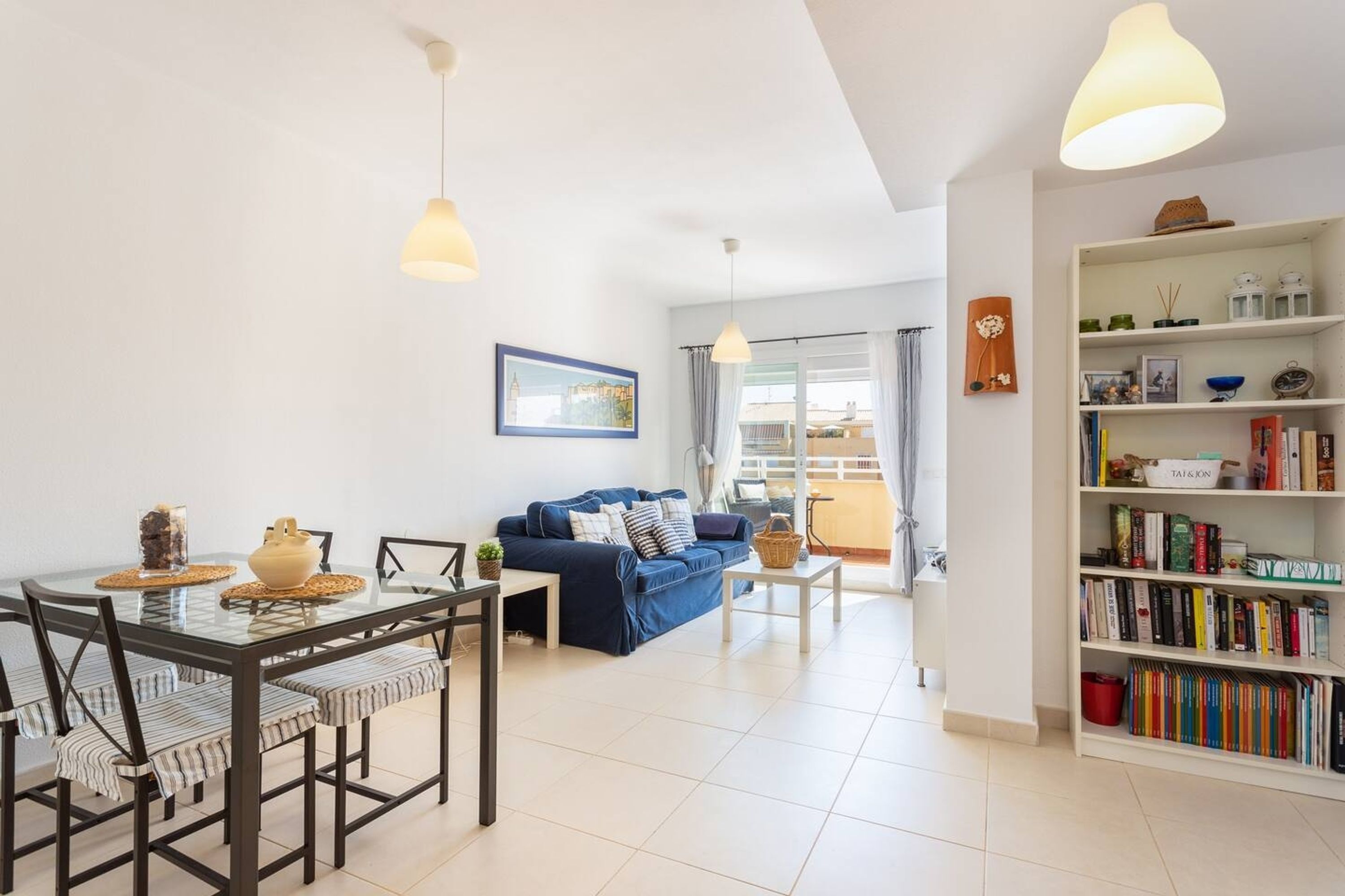 A modern living room with a glass dining table, blue sofa, and a bookshelf filled with books.