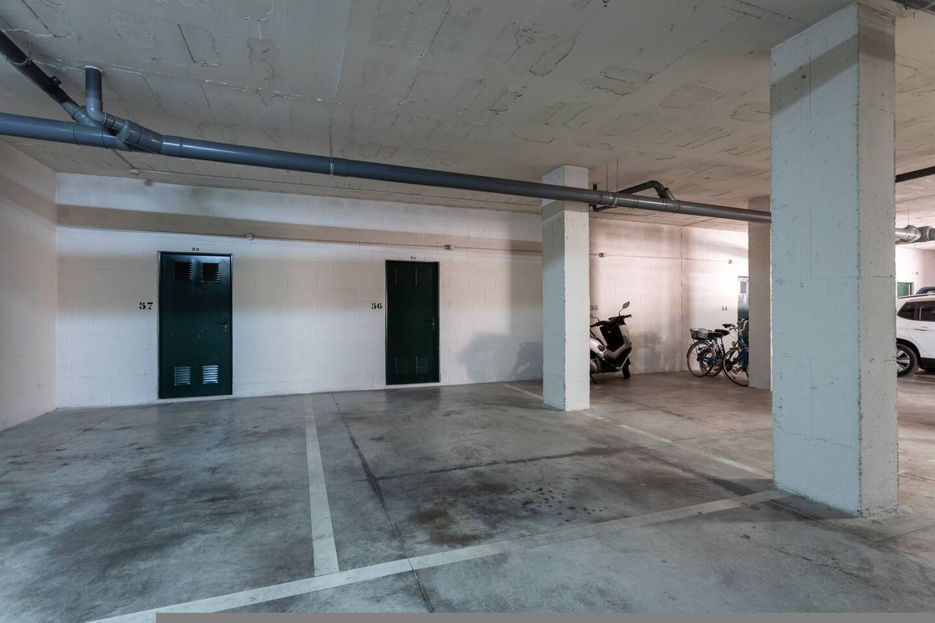 Un estacionamiento con pisos de concreto, dos puertas con los números 57 y 56, y una fila de bicicletas estacionadas.