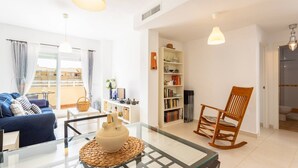 A modern living room with a sofa, a glass coffee table, a wooden rocking chair, a bookshelf, and a view of the outside. - Duplex Algarrobo Costa Solset (Algarrobo)