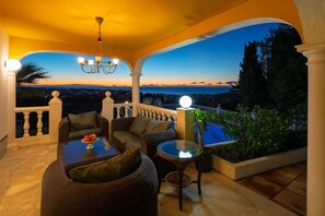 Un espace extérieur abrité, aménagé avec des meubles en osier, une table en verre et offrant une vue sur la mer au crépuscule.