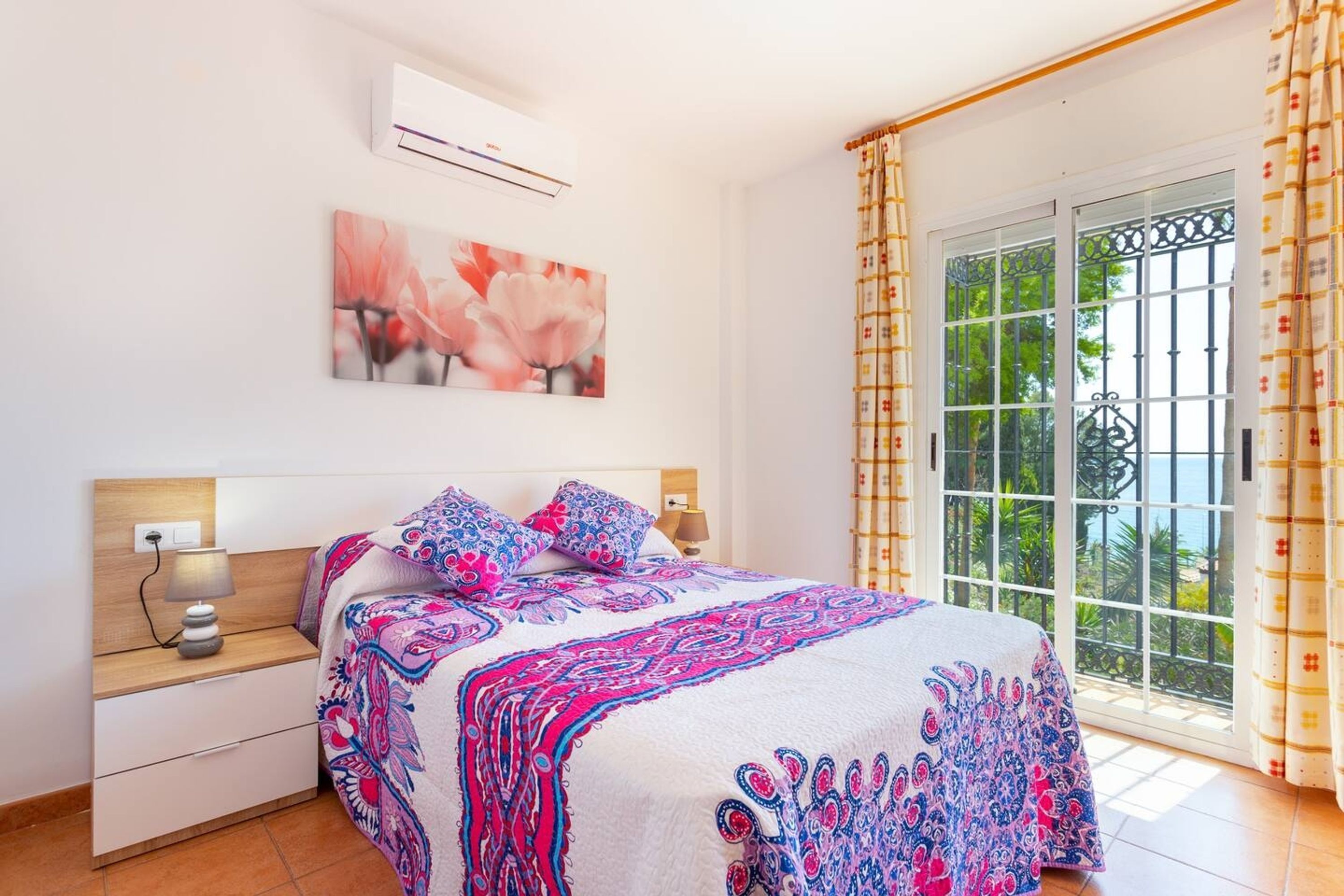 A bedroom with a bed, nightstand, lamp, air conditioning unit, and a window with curtains.