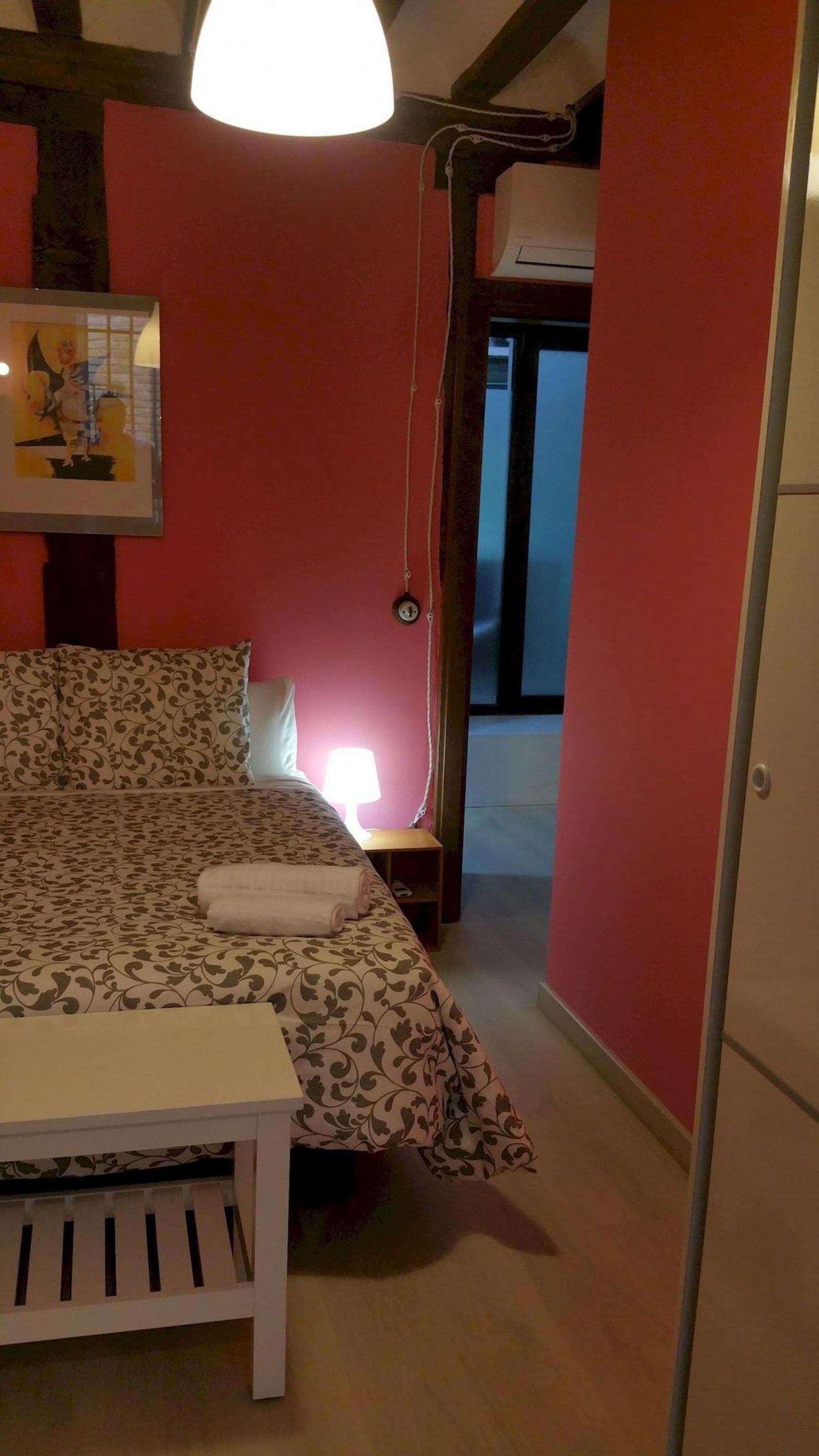 Een kamer met een rode muur, een raam, een bed met beddengoed met een patroon en een witte stoel.