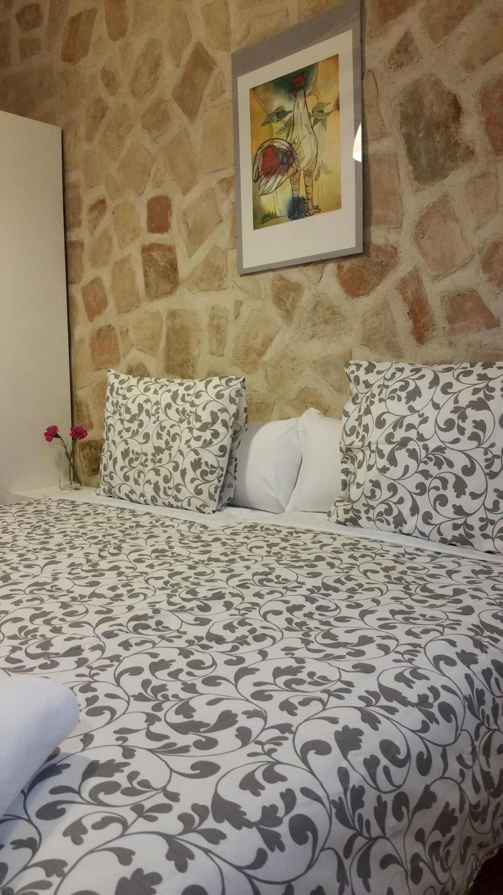 Una habitación con pared de piedra, una obra de arte enmarcada, una cama con ropa de cama estampada y un florero con una sola flor.