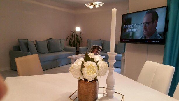 A modern living room with a white sofa, a coffee table with a vase of white flowers, and a flat-screen TV mounted on the wall. - Apartamento Deluxe 2 Cason DEL Corpus 2 Go Native Toledo 11 Plazas (Toledo)