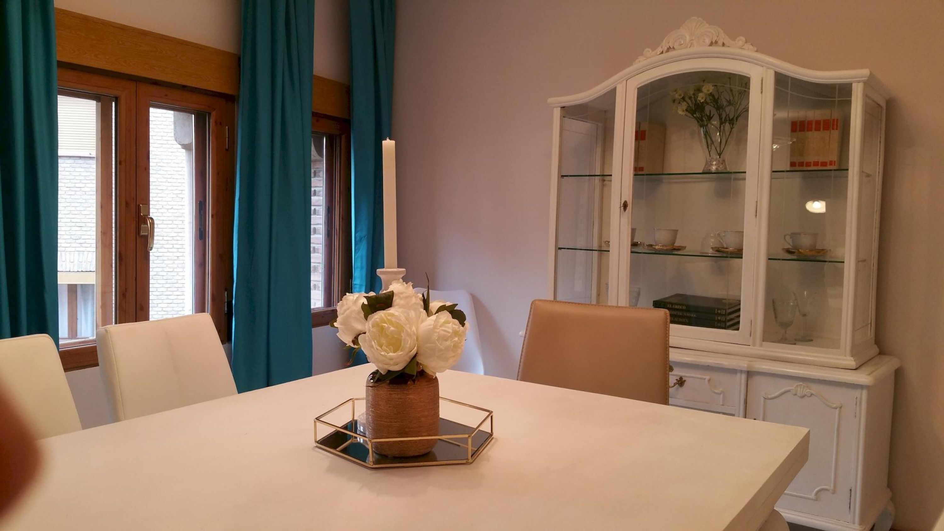 Un coin repas avec une table blanche, des chaises, un vase de fleurs et un buffet vitré contenant de la vaisselle.