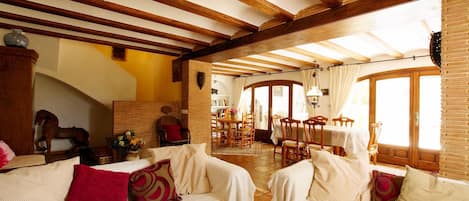 A spacious living area with high ceilings and exposed wooden beams, featuring two white sofas with red cushions, a dining area with wooden furniture, and large windows allowing natural light.