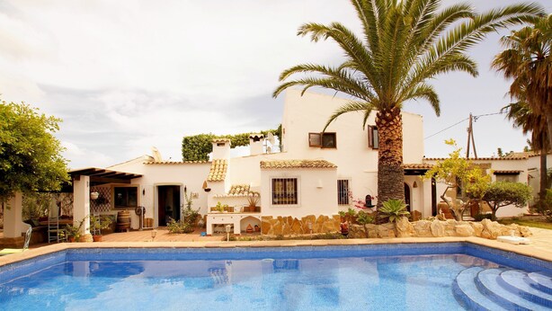 A swimming pool with a tiled edge, surrounded by a white building with a tiled roof, palm trees, and a stone wall. - Villa La Siesta in Javea (Javea)