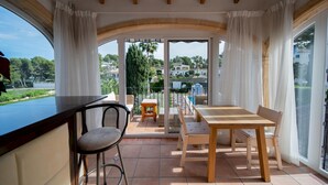 Een zonnig terras met een houten tafel en stoelen, een bar en uitzicht op een weelderige tuin en gebouwen door grote glazen deuren.