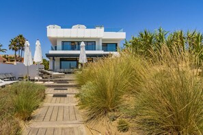 A modern building with a wooden deck leading to a pool area, surrounded by tall grasses and palm trees. - Villa Frontline Beach Costabella - Marbella (Marbella)