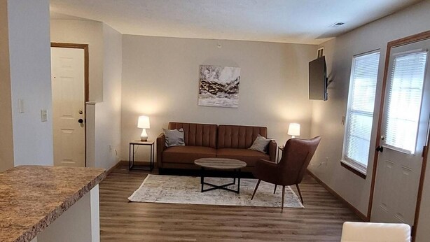 A modern apartment with a kitchen, dining area, and living room. The kitchen has a granite countertop and a built-in oven. The dining area has a table and chairs. The living room has a couch and a coffee table. There is a painting on the wall and a window with blinds. - 2 Bedroom Apartment Next To Rivian (Bloomington)