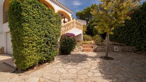 A courtyard with stone flooring, a white building with arches, and a well-maintained hedge.