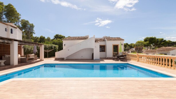 A swimming pool with a white building in the background, a pergola with furniture, and a clear sky.