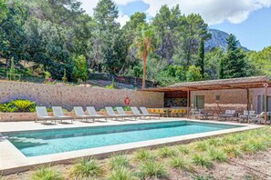 A pool area with lounge chairs and a covered seating area, surrounded by trees and a mountainous backdrop. - Finca Val de Marc in Pollen a (Pollença)