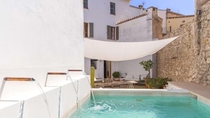 A pool area with a stone wall, a white building, and a shaded seating area. - Casa Campomar by Vida Villas (Pollença)