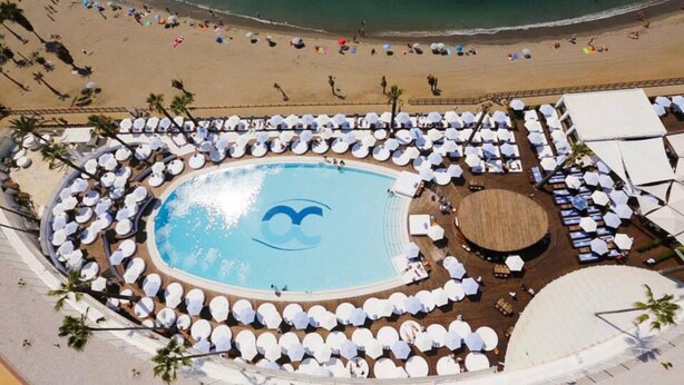 Vue aérienne d’un complexe hôtelier doté d’une piscine centrale, de chaises longues et d’une zone de plage.