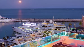 Un port de plaisance où sont amarrés de nombreux yachts, un espace piscine abrité par des parois de verre et un bâtiment doté d’un toit de chaume.