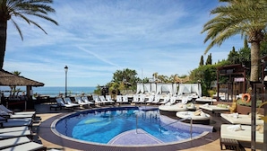 Une piscine circulaire entourée de chaises longues et de parasols, bordée de palmiers et offrant une vue sur l’océan.