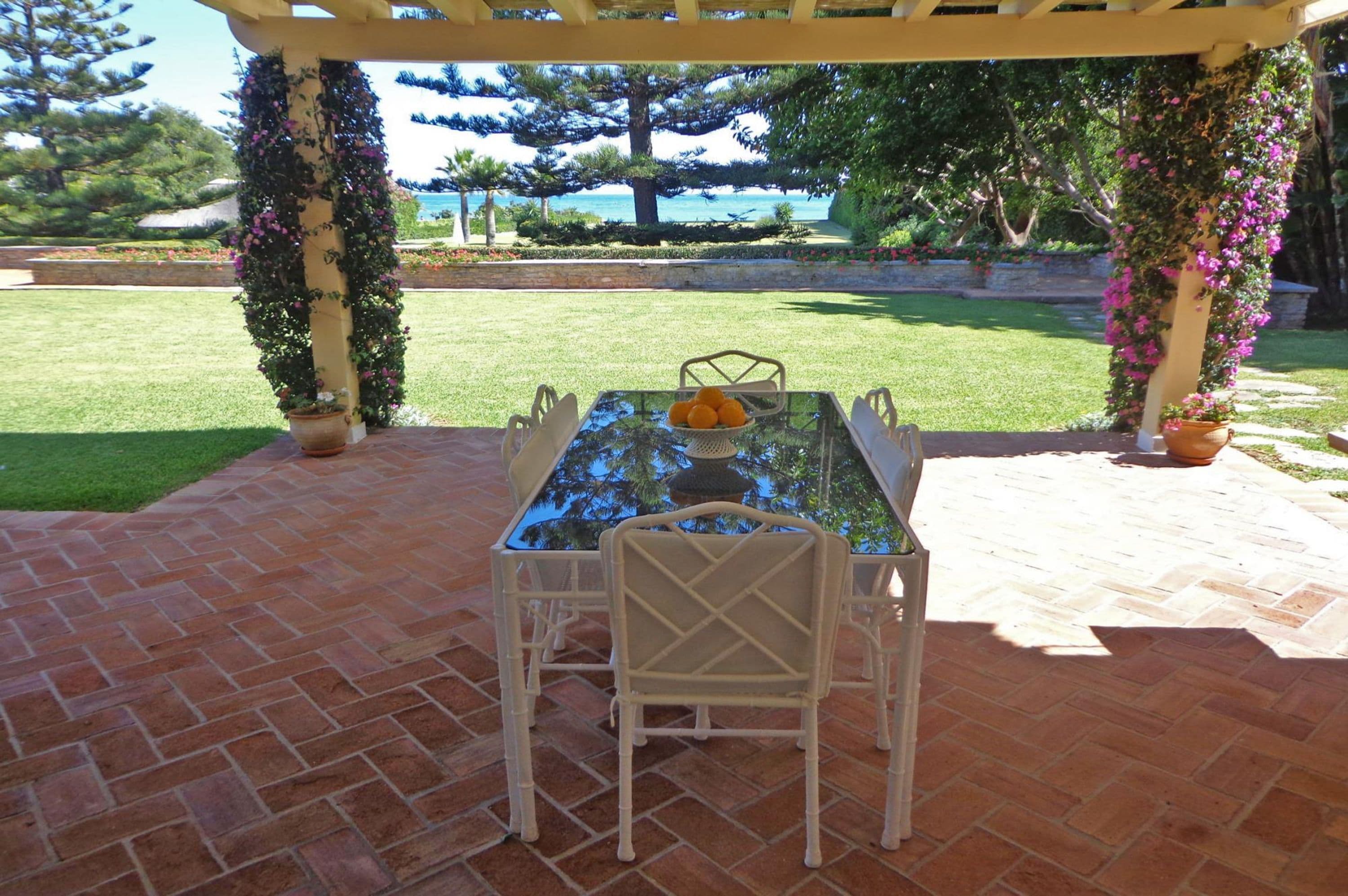A covered patio with a table and chairs, surrounded by potted plants and a view of the sea.