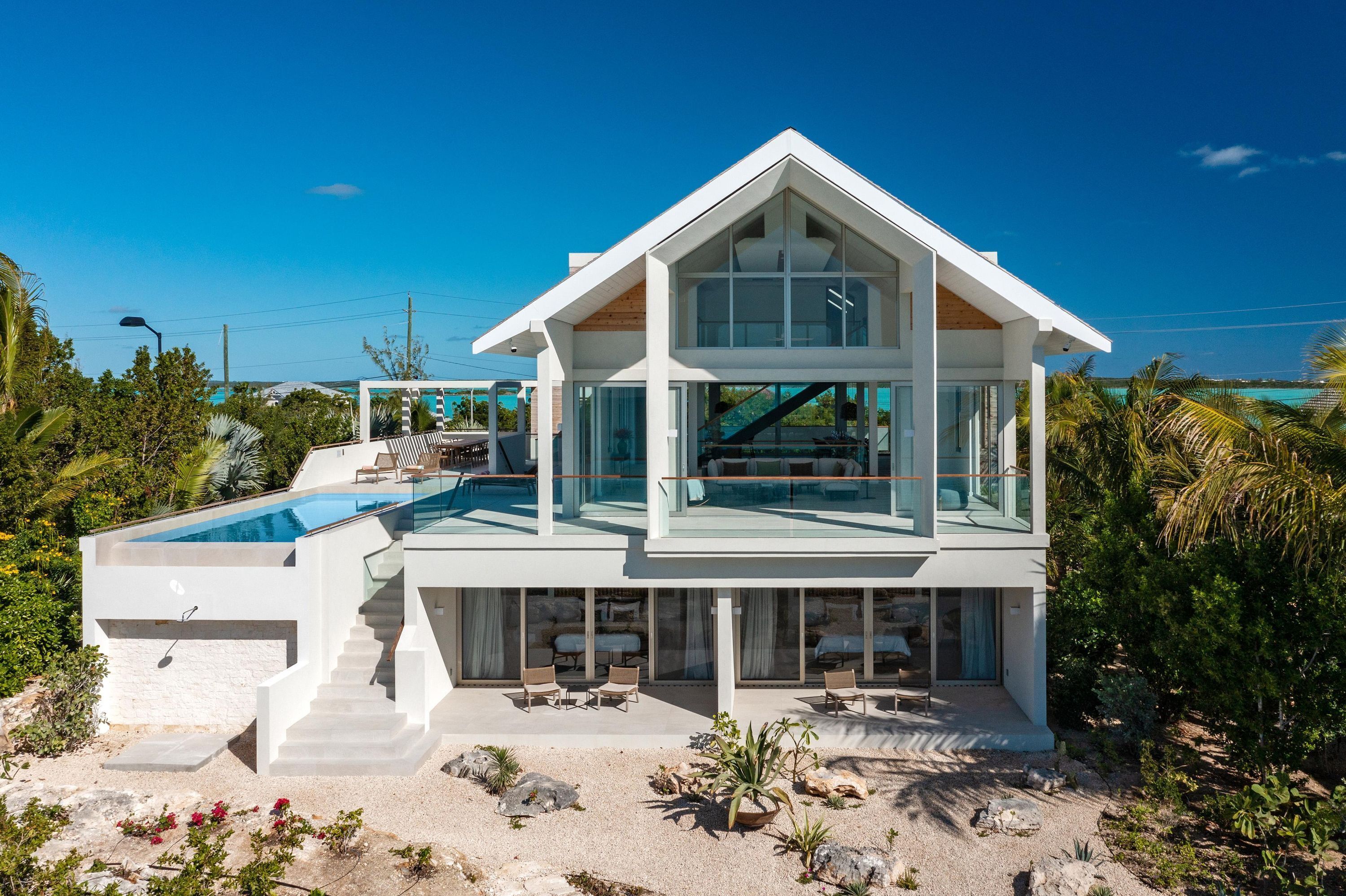 A modern two-story house with a glass-enclosed swimming pool, outdoor seating, and a view of the ocean.