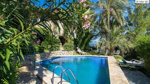 A clear blue pool surrounded by lush greenery and palm trees, with a stone wall and lounge chairs. - El Vilar 60 Villa Puerto Pollenca (Pollença)
