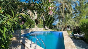 A clear blue pool surrounded by lush greenery and palm trees, with a stone wall and lounge chairs. - El Vilar 60 Villa Puerto Pollenca (Pollença)