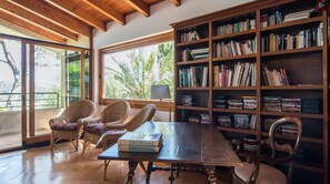 A room with a large bookshelf, wicker chairs, a table with books, and a view through large windows. - El Vilar 60 Villa Puerto Pollenca (Pollença)