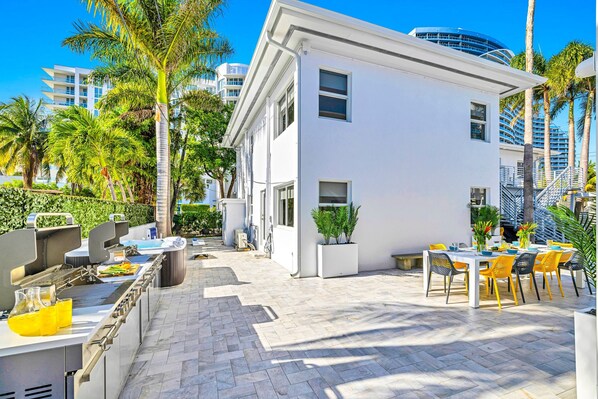 Un patio exterior moderno con una parrilla, mesa para comer y sillas, rodeado de palmeras y un edificio blanco.