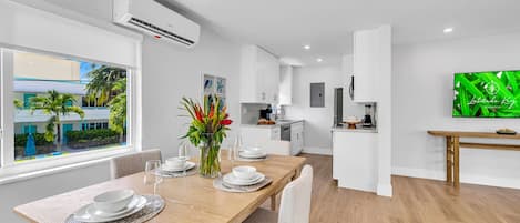 A modern kitchen with white cabinets, a dining area with a wooden table and chairs, and a TV mounted on the wall.