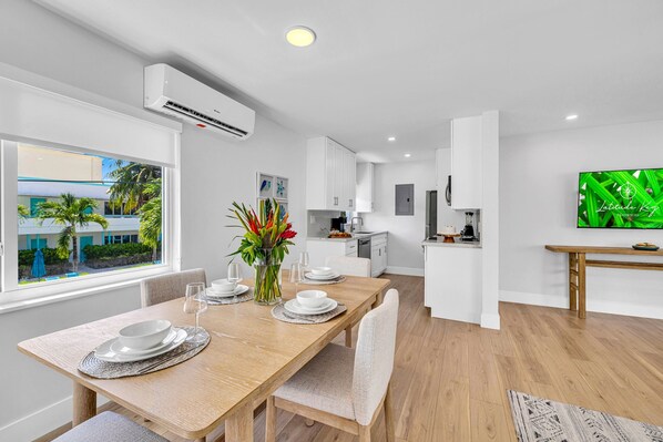 A modern kitchen with white cabinets, a dining area with a wooden table and chairs, and a TV mounted on the wall.