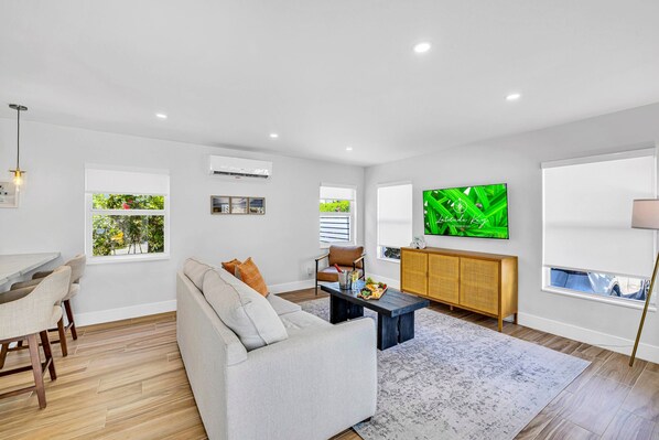 A modern living room with a sofa, coffee table, dining area, and a TV on the wall.
