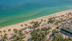 Vue aérienne d’une plage bordée de palmiers, d’un parking et d’un bâtiment doté d’un toit plat.
