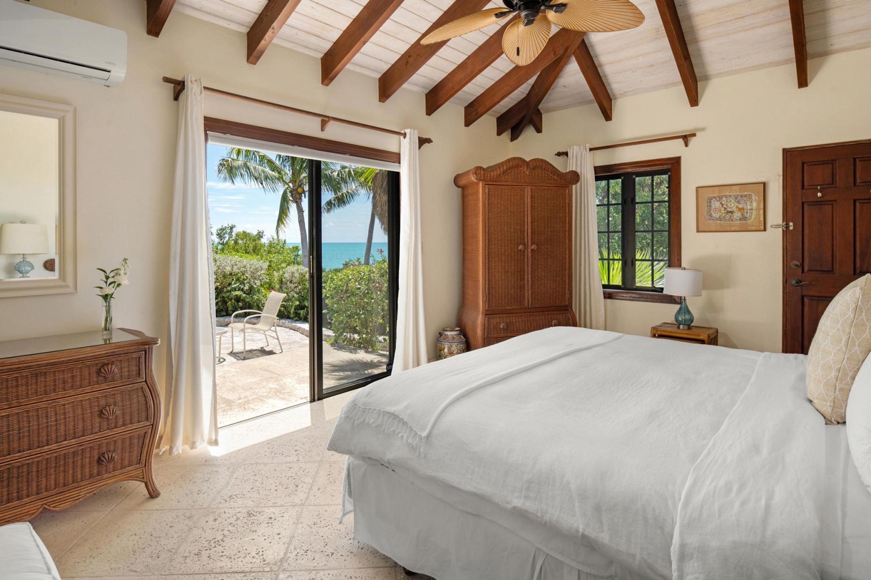 A bedroom with a bed, a wooden wardrobe, a nightstand, and a window with a view of palm trees and the ocean.