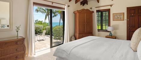 A bedroom with a bed, a wooden wardrobe, a nightstand, and a window with a view of palm trees and the ocean.