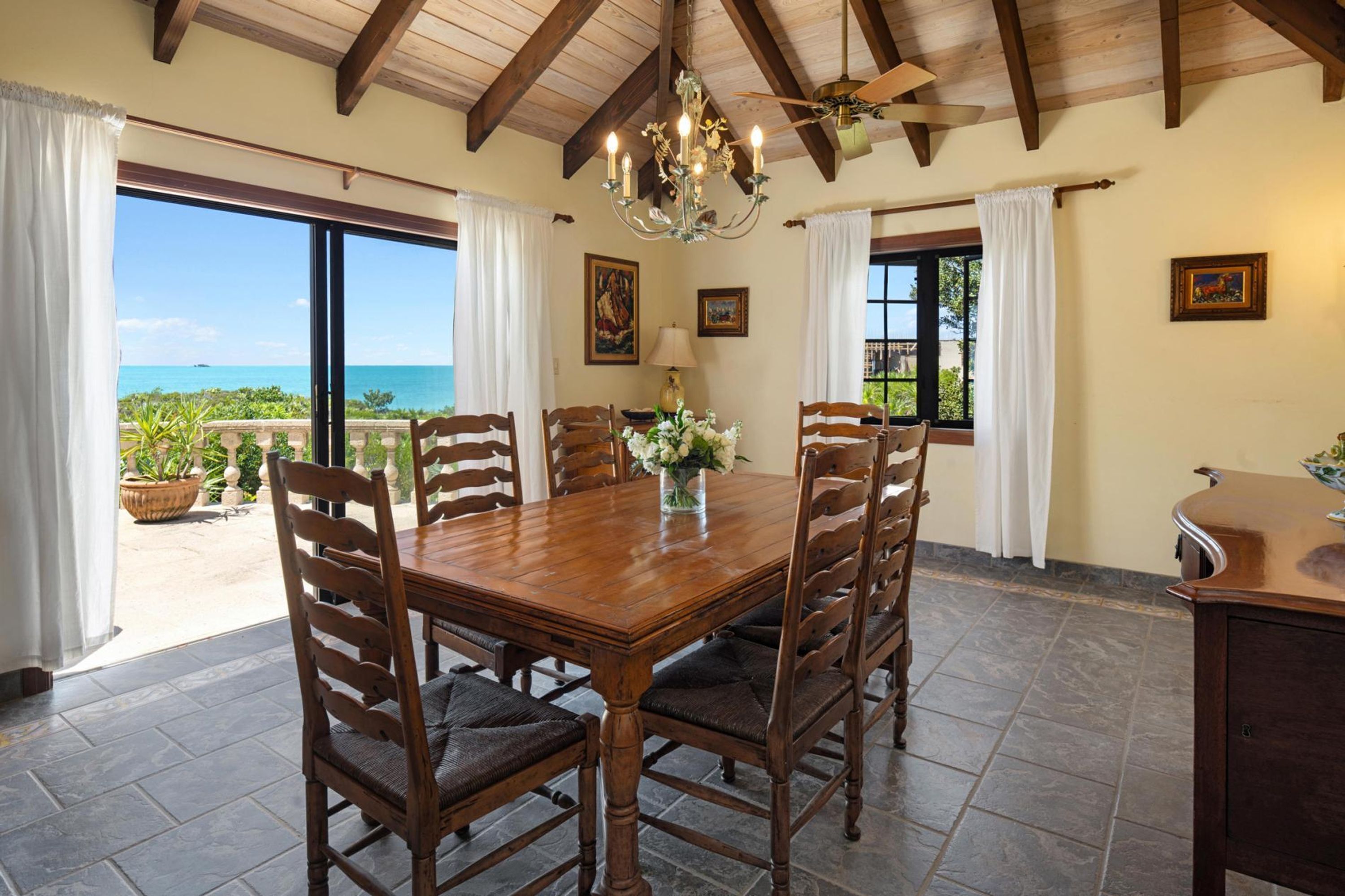 A dining area with a wooden table and chairs, a chandelier, and a view of the ocean.