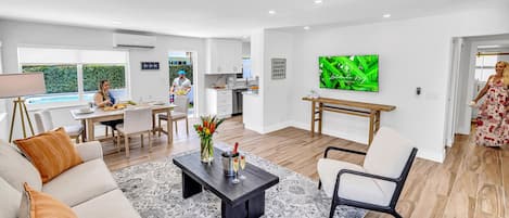 Una sala de estar moderna con un sofá, una mesa de centro y un televisor. Hay un comedor con una mesa y sillas, y una cocina al fondo.