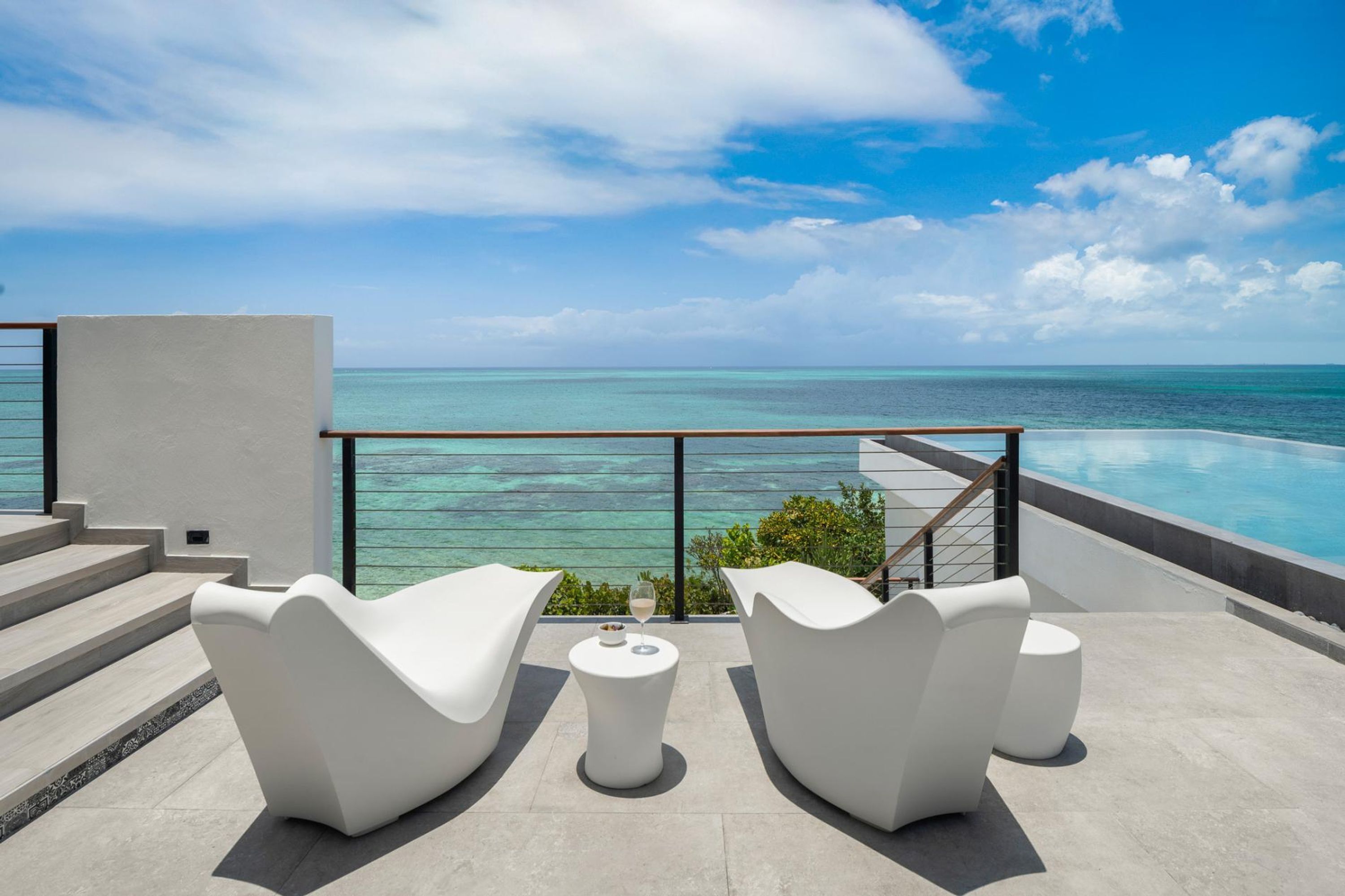 Ein moderner Außenbereich mit weißen Stühlen und einem kleinen Tisch, mit Blick auf das klare, blaue Meer und einen Swimmingpool.