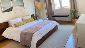 A neatly arranged bedroom with a bed, bedside table, a mirror, a window with curtains, and a potted plant. - Spacious Glyfada Golf 4BR -near Beach Shops (Glyfada)