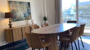 A modern dining area with a wooden table and chairs, a large painting on the wall, and a view of buildings through the window. - Spacious Glyfada Golf 4BR -near Beach Shops (Glyfada)