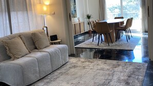 A modern living room with a sofa, a dining table, and a kitchen area visible through an open doorway. - Spacious Glyfada Golf 4BR -near Beach Shops (Glyfada)