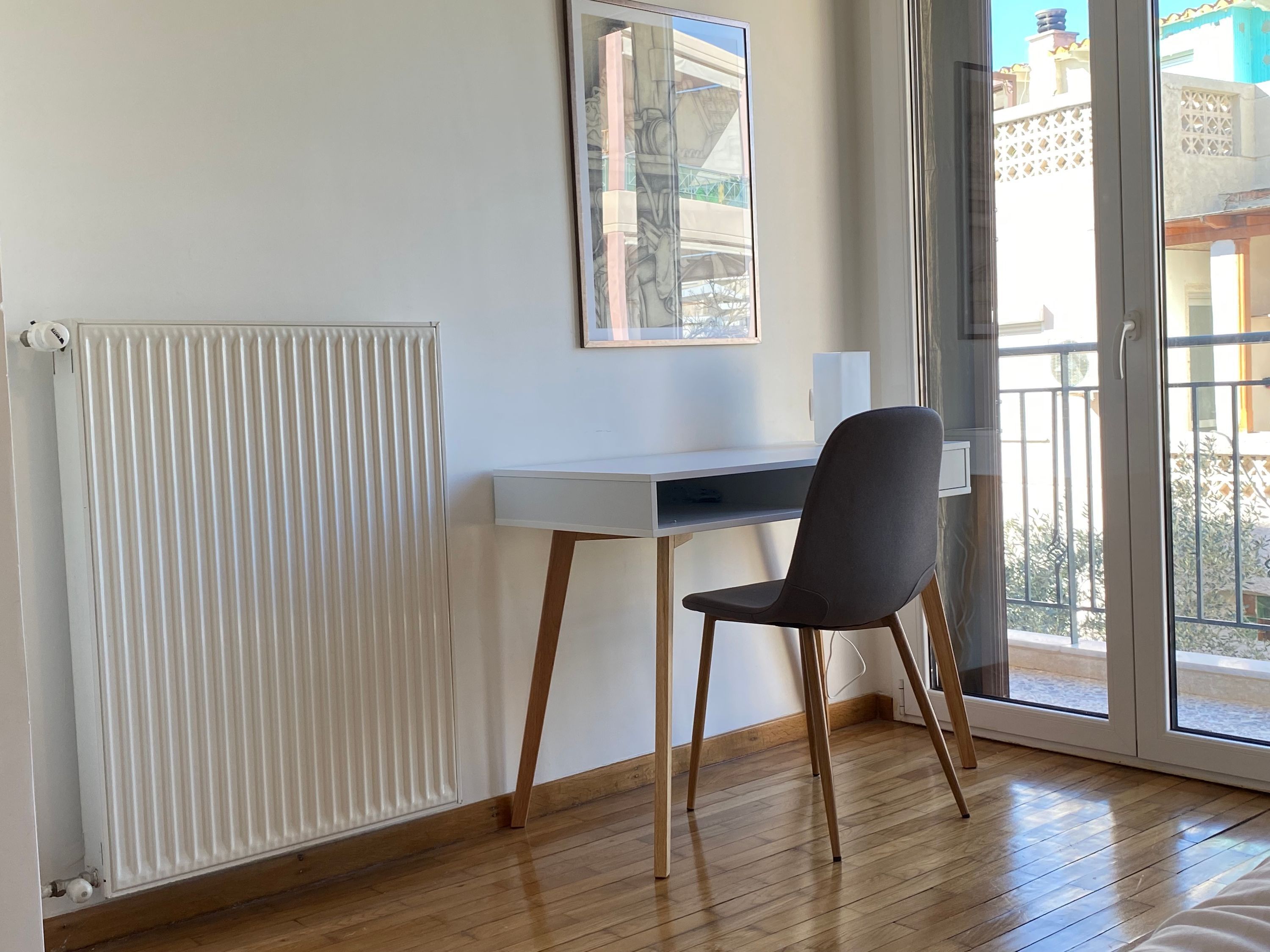 A room with a white desk, a grey chair, a radiator, and a window with a view of buildings.