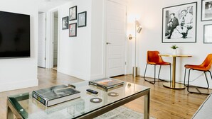 A modern living room with a glass coffee table, a flat-screen TV, and a dining area with two orange chairs and a small table.