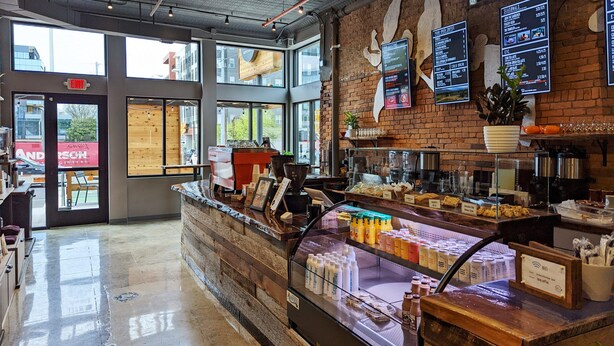 A coffee shop interior with a counter displaying various beverages, a menu board, and a brick wall.