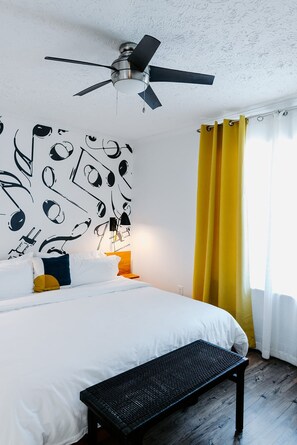 A bedroom with a bed, a ceiling fan, a wall with musical notes, a bench, and yellow curtains. - The Vinyl at Music Row - 1 Bedroom Apartment (Nashville)