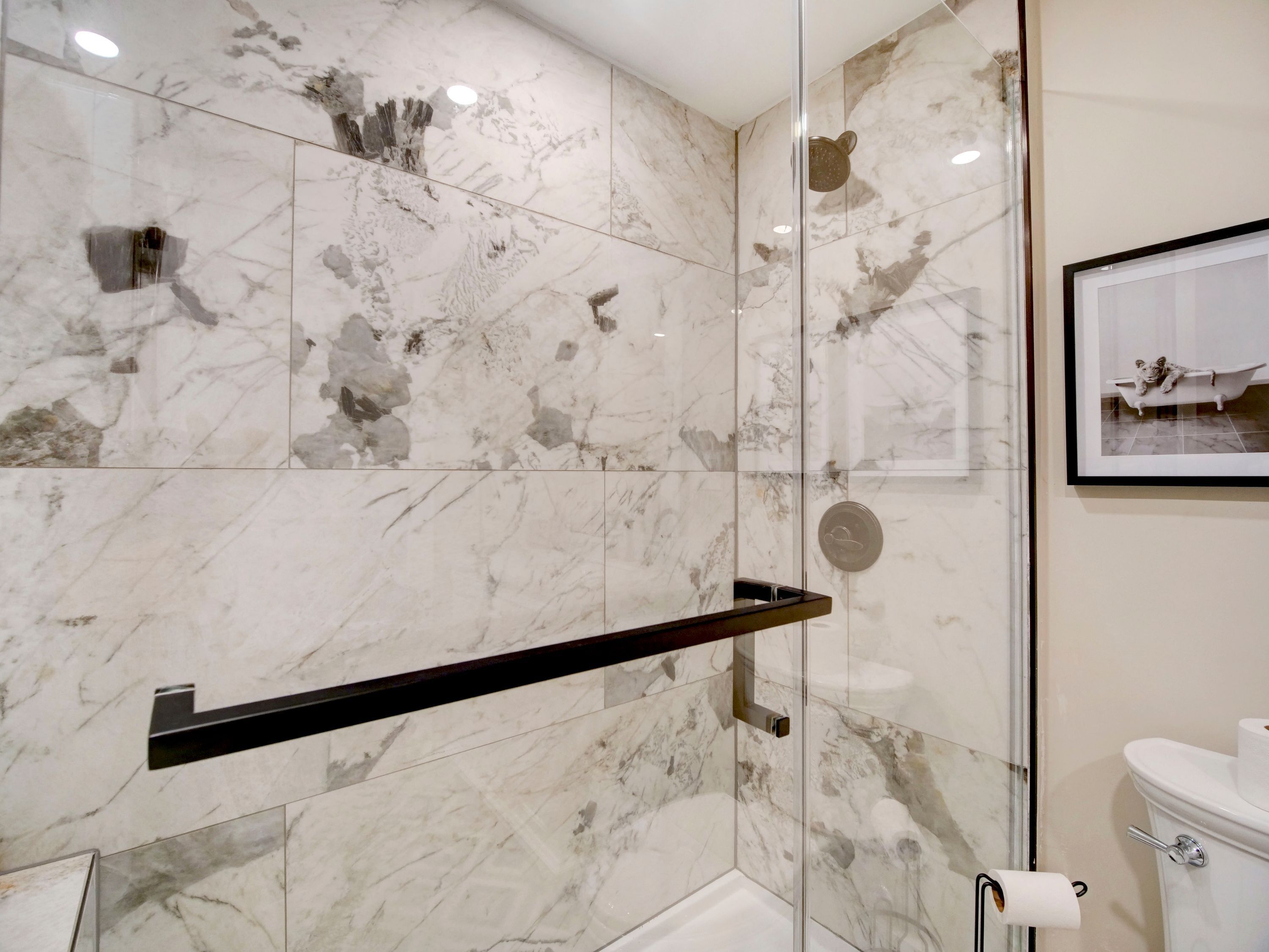 Un baño con paredes de mármol, cabina de ducha de vidrio, toallero negro y un cuadro en la pared.