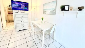 A modern, white-themed room with a flat-screen TV, a dining table with chairs, a white cabinet with drawers, a painting on the wall, and a small shelf with decorative items. - Southwinds 2 for 3 King Studio for 3 w Kitchen (Hollywood)