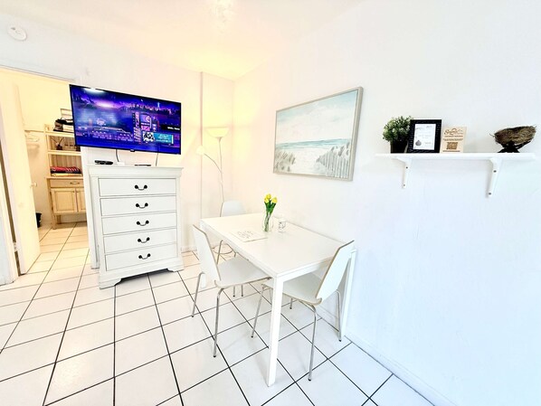 A modern, white-themed room with a flat-screen TV, a dining table with chairs, a white cabinet with drawers, a painting on the wall, and a small shelf with decorative items. - Southwinds 2 for 3 King Studio for 3 w Kitchen (Hollywood)