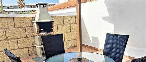 En täckt uteplats med ett runt bord i glas och svarta flätade stolar, en grill och utsikt över byggnader i bakgrunden.