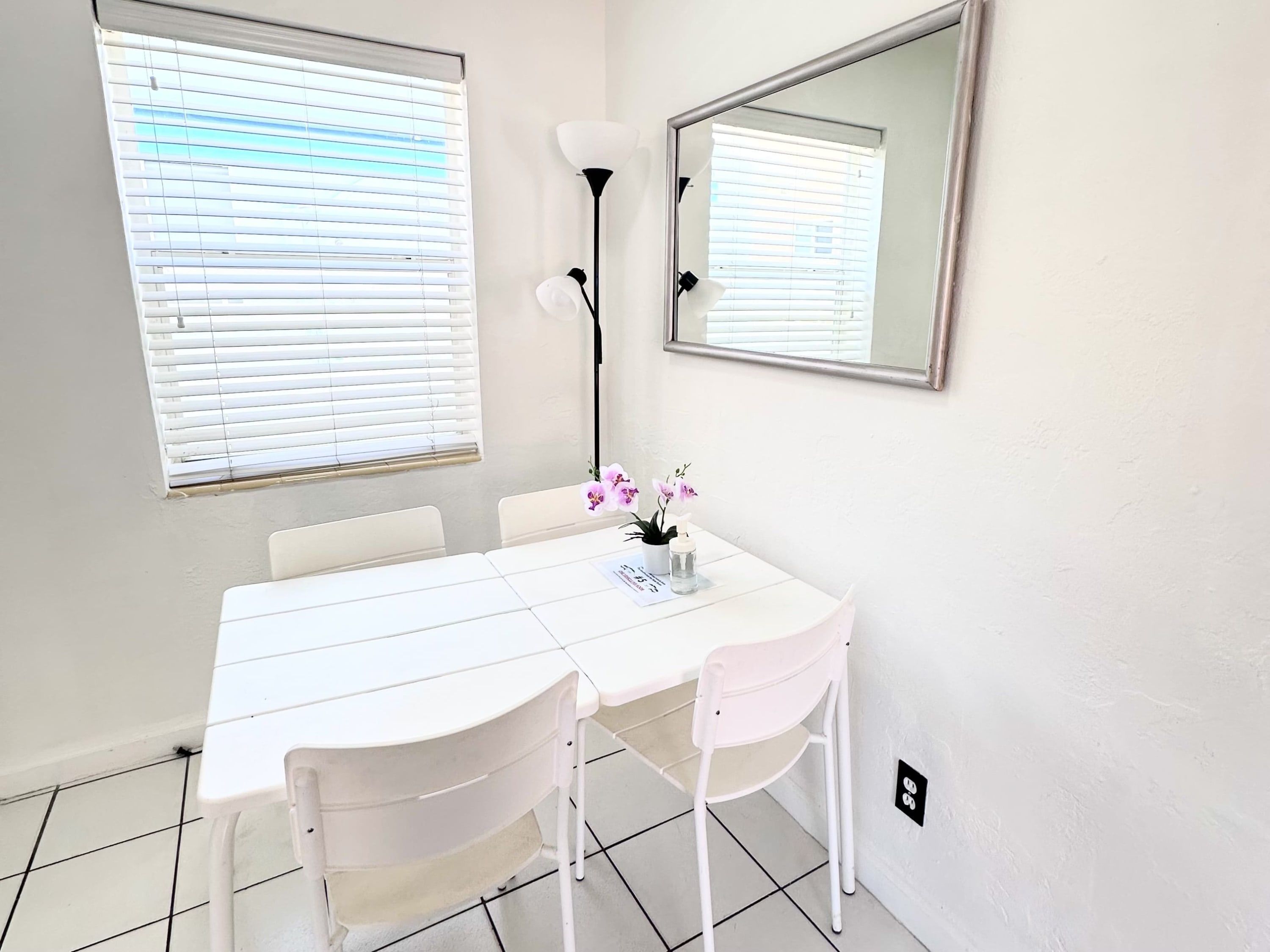 A white dining area with a table, chairs, a mirror, and a window with blinds.